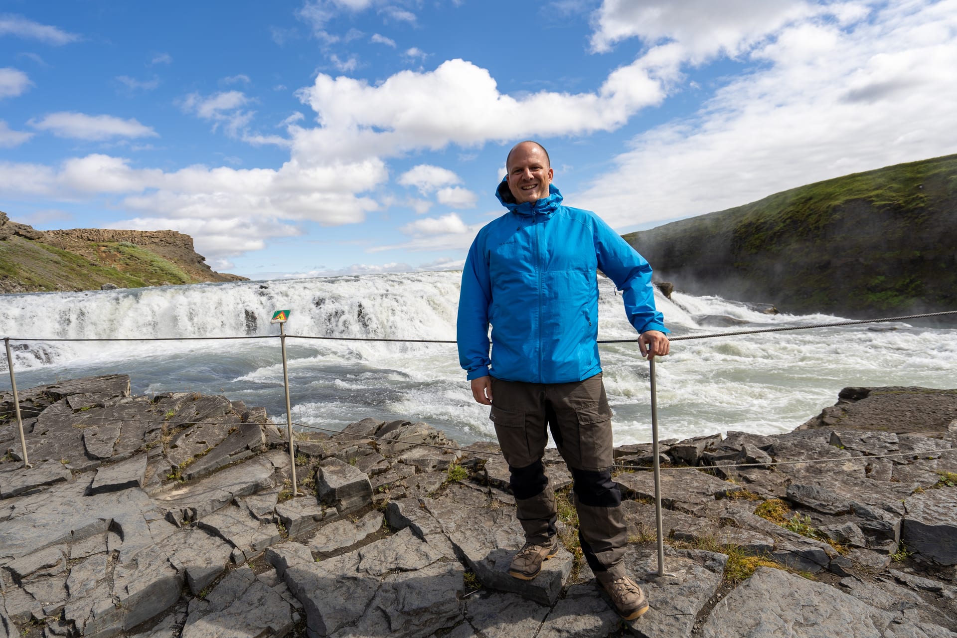 Mann in blauer Jacke vor dem mächtigen Gullfoss-Wasserfall in Island, dahinter tosende Stromschnellen und grüne Schlucht.