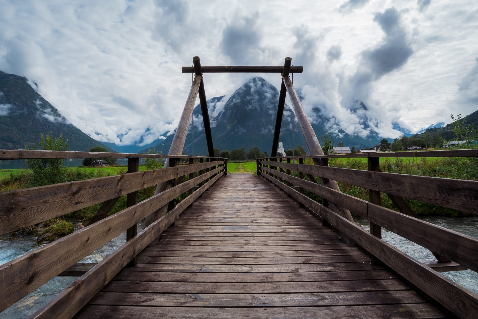 Hölzerne Hängebrücke über einen Fluss, im Hintergrund wolkenverhangene Berge und ein Fjordtal in Norwegen.